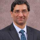 This image is a headshot of a person wearing glasses, a dark suit, a striped shirt, and a blue tie, set against a light brown background.