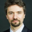Headshot of a person with short brown hair and a beard, wearing a dark suit, light shirt, and tie, set against a dark background.