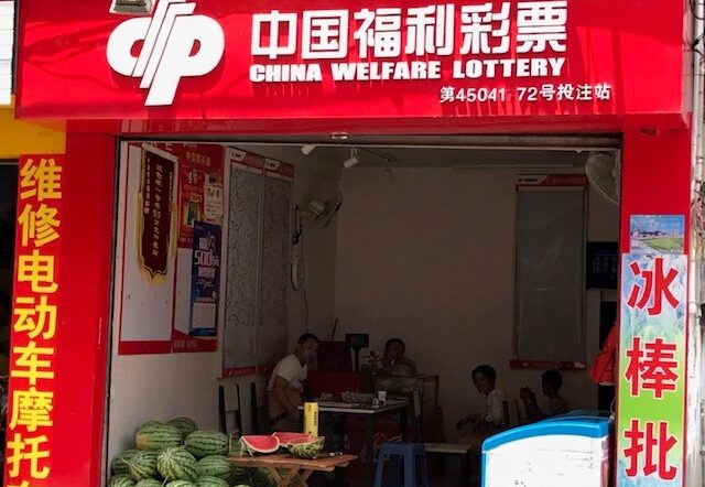 An outdoor view of a "China Welfare Lottery" store. The storefront is red with Chinese characters and logos, while watermelons are displayed at the entrance. Several people are inside the shop.