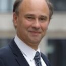 Headshot of a person in formal attire, wearing a suit and tie, with a slight smile, in an outdoor setting with a blurred background.