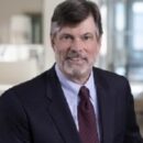 A professional headshot of a person wearing a suit and tie, with a blurred office background.
