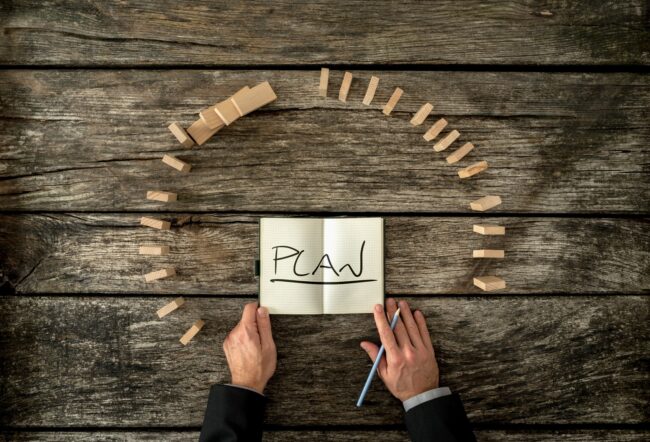 A pair of hands holds an open notebook with the word "PLAN" written on it, surrounded by a circular arrangement of falling wooden blocks on a wooden surface.
