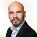 Headshot of a person with a bald head and a beard, wearing a dark suit jacket and a light blue collared shirt, smiling slightly against a plain background.