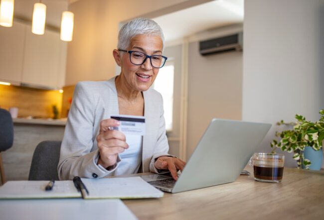A person with short gray hair and glasses is smiling, holding a credit card while using a laptop. A glass of coffee is on the table in a cozy room.