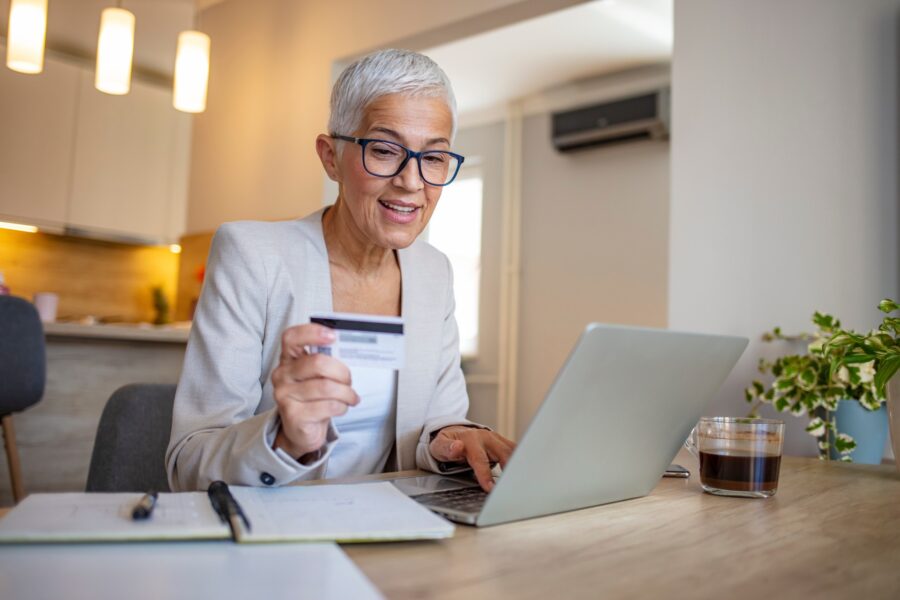 A person with short gray hair and glasses is smiling, holding a credit card while using a laptop. A glass of coffee is on the table in a cozy room.