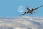 A commercial airplane flying above the clouds with a large moon in the background against a clear blue sky.