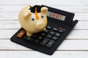 A piggy bank wearing a graduation cap sits on a calculator, with the word "DEBT" displayed. This represents student loan debt.