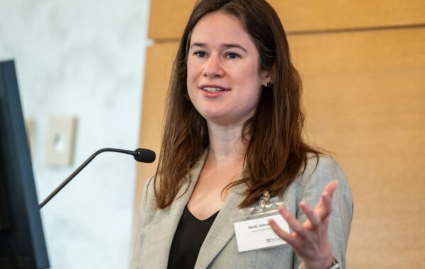 A person in a suit giving a presentation, holding a remote and speaking into a microphone with a screen nearby.