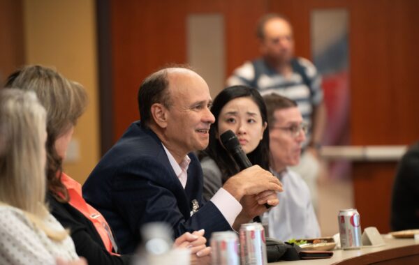 A group of people in a conference or meeting setting, with one person speaking into a microphone. The table has drinks and snacks, and the background shows a blurred figure.