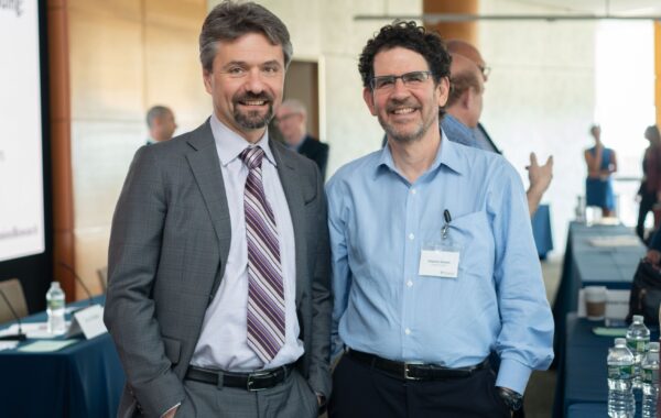 Two people standing side by side in a conference setting, one wearing a suit and tie and the other in a blue shirt with a name badge.