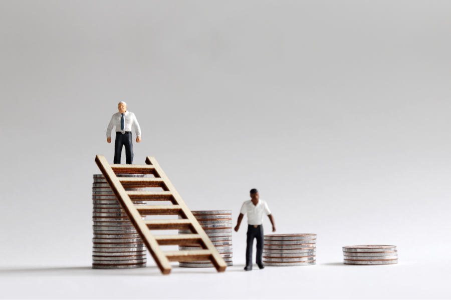 The image shows two miniature figures in business attire standing on stacks of coins of varying heights. A wooden ladder connects two of the stacks. This represents economic disparity or inequality.