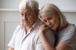 An older couple in an emotional moment, with one person comforting the other by resting their head on the other's shoulder.