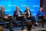 A panel of four individuals seated and engaged in discussion, with a blue backdrop displaying the word "Brookings."