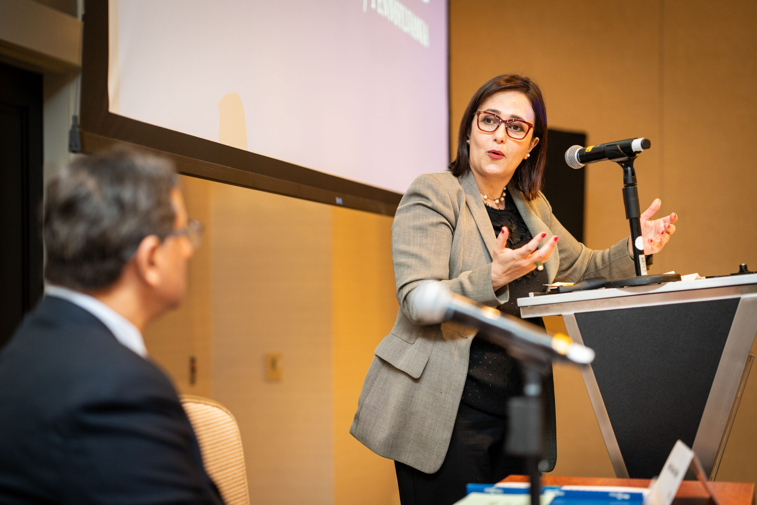 A person in a suit speaks at a podium with a microphone, engaging an audience, while another person listens. A projection screen is visible in the background.