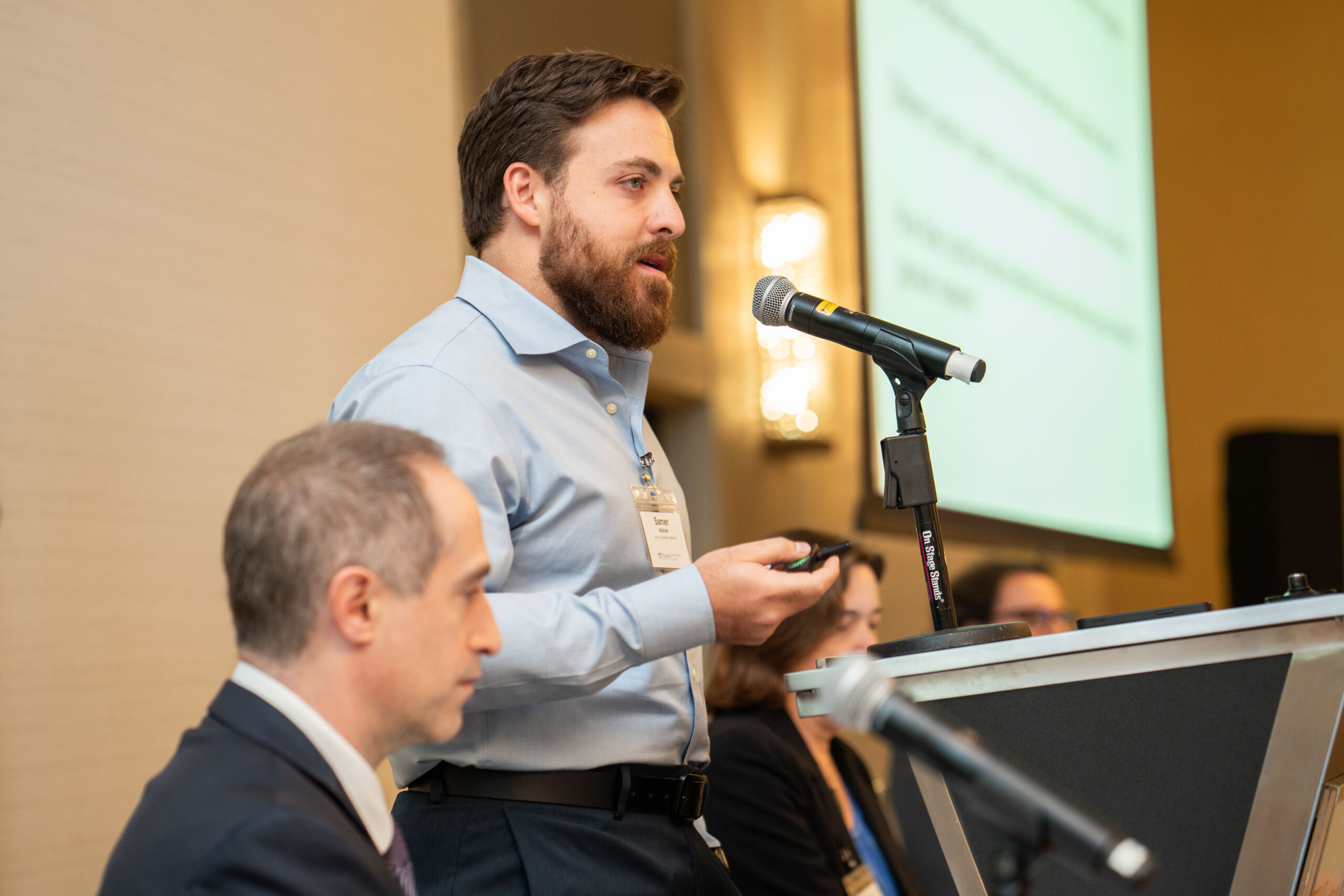 A person speaking at a podium with a microphone during a conference or meeting. Other people are seated nearby, and a presentation screen is visible in the background.