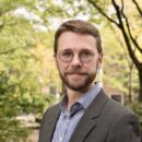 Headshot of a person with glasses and a beard, wearing a suit jacket and checked shirt, standing outdoors with trees in the background.