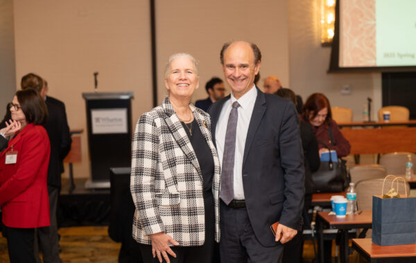 A smiling couple is pictured at a formal event, with the person on the left wearing a checkered jacket, and the person on the right in a suit. Other attendees are in the background.
