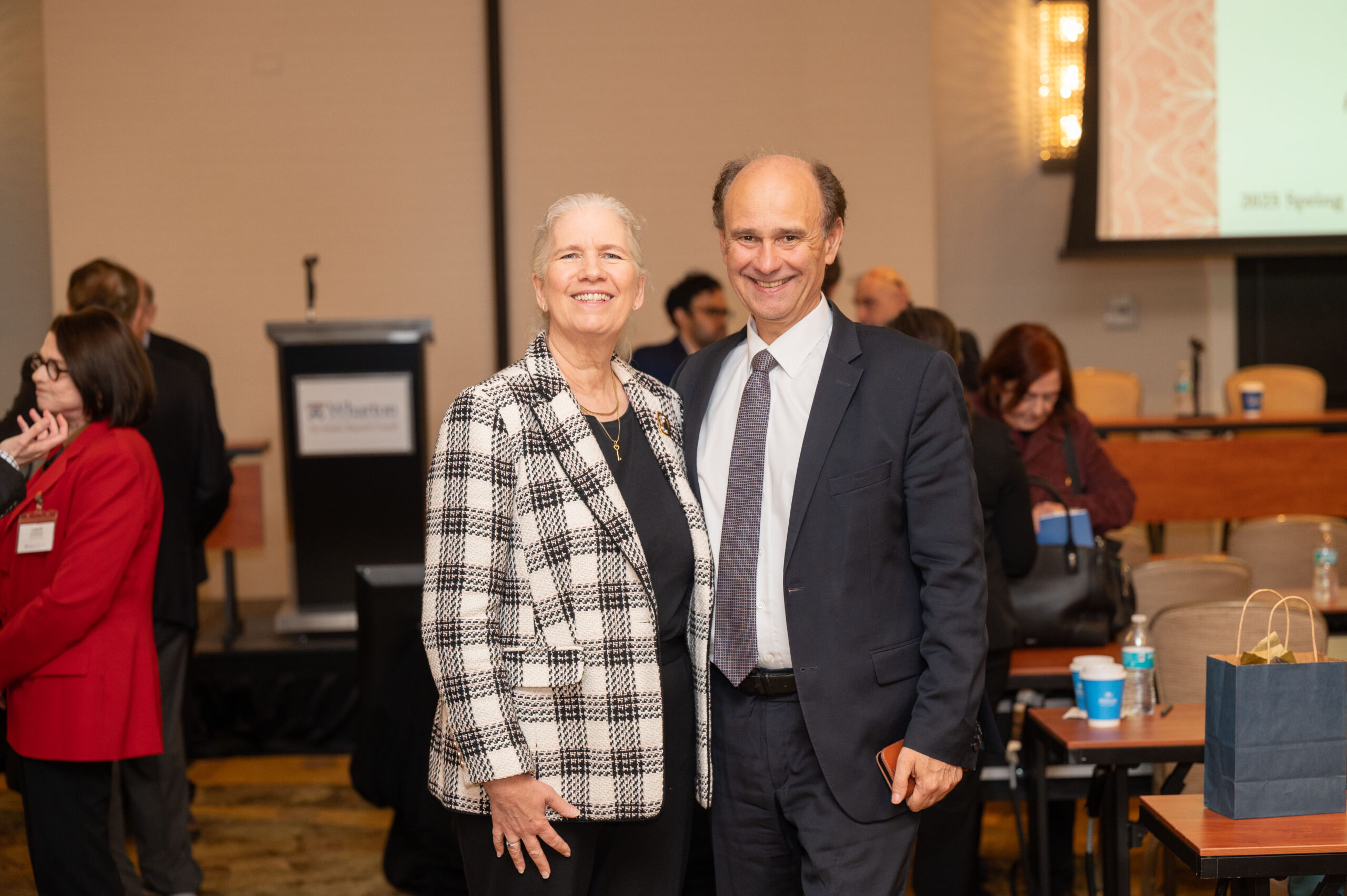 A smiling couple is pictured at a formal event, with the person on the left wearing a checkered jacket, and the person on the right in a suit. Other attendees are in the background.
