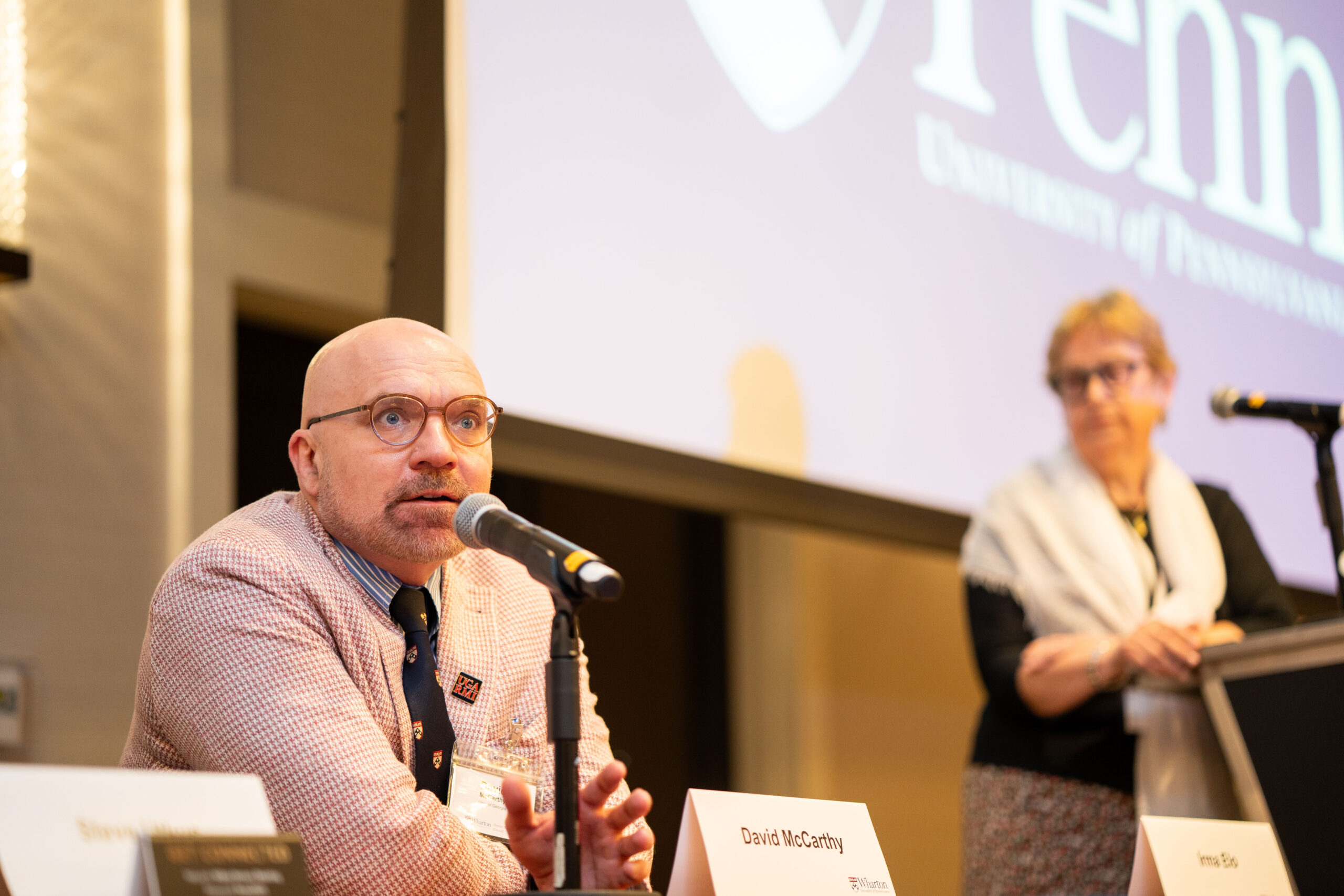 A person speaking into a microphone at a panel event, with another person standing in the background near a podium. The setting appears to be academic or professional, suggested by the screen displaying part of a university logo.