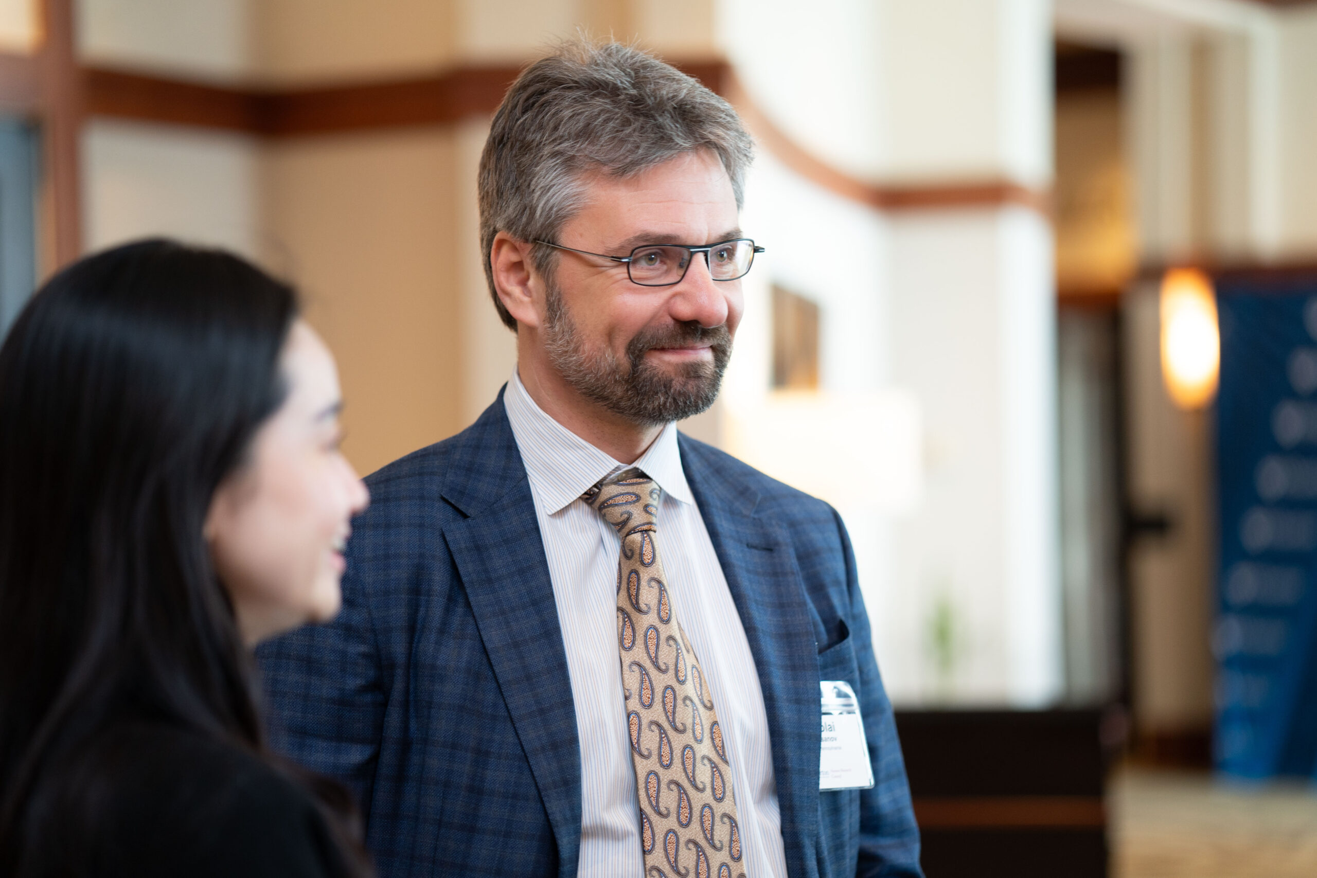 A couple of people in business attire are engaged in conversation at an indoor event, with one wearing glasses and a suit with a patterned tie.