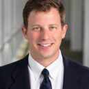 A person wearing a suit with a white shirt and a blue polka dot tie, smiling in a professional setting.