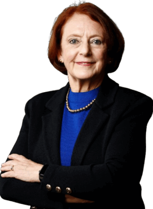 A person with short brown hair wearing a black blazer and blue top, arms crossed. They are looking forward with a confident expression.